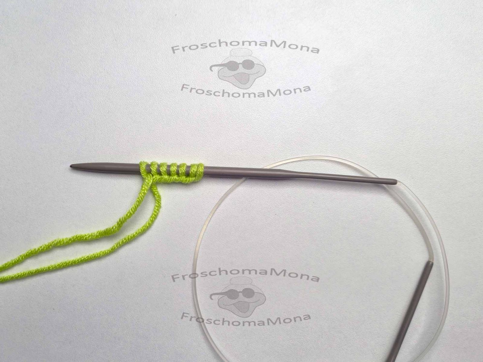 Auf einer weißen Unterlage liegt eine Rundstricknadel auf der bereits 7 Maschen mit grünem Garn angeschlagen wurden.