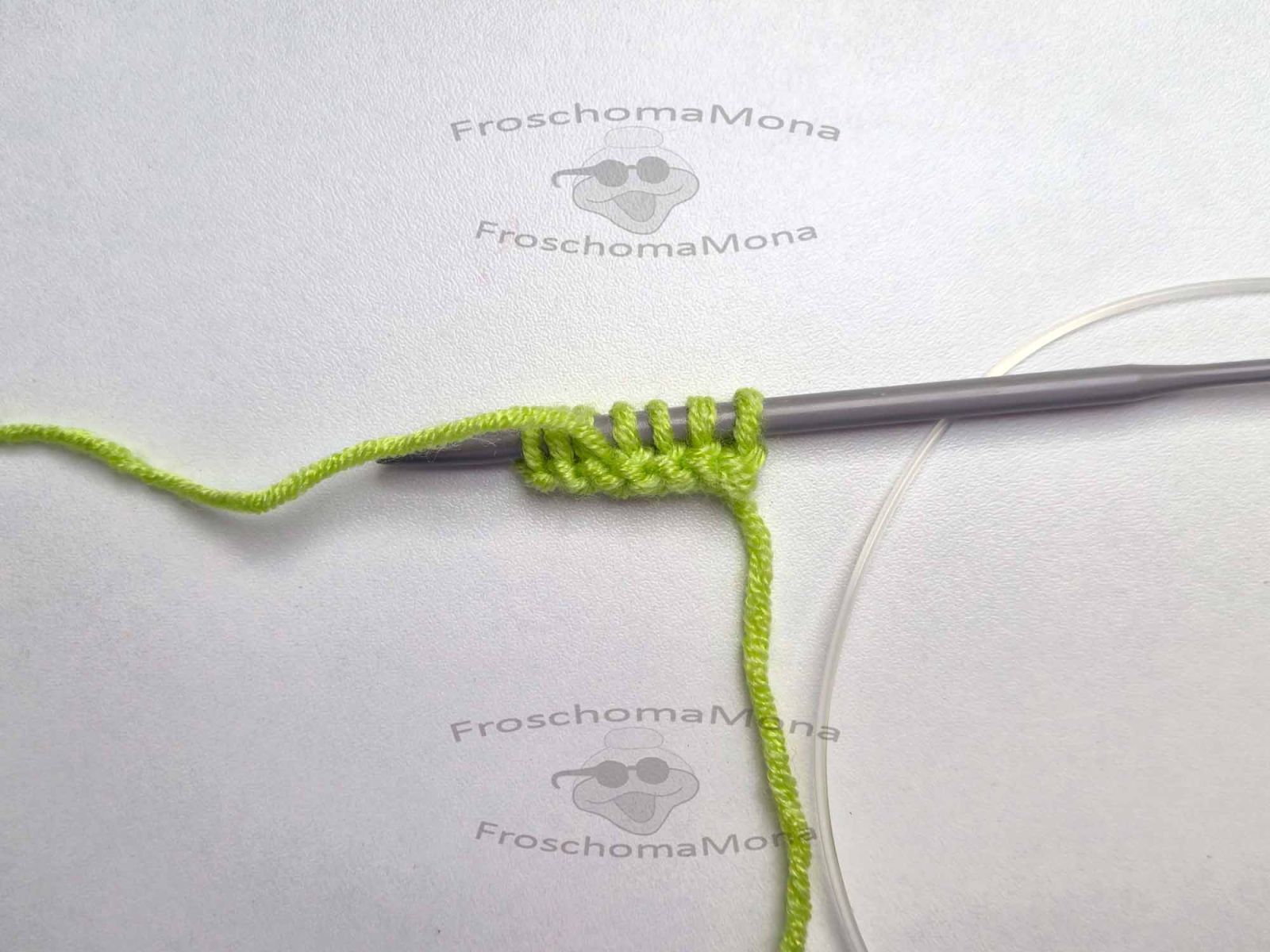 Die Stricknadel auf weißem Untergrund zeigt das Tuch Dana nach der 2. Reihe. Der Arbeitsfaden liegt vor den 3 Maschen des I-Cord-Randes am Ende der Reihe.