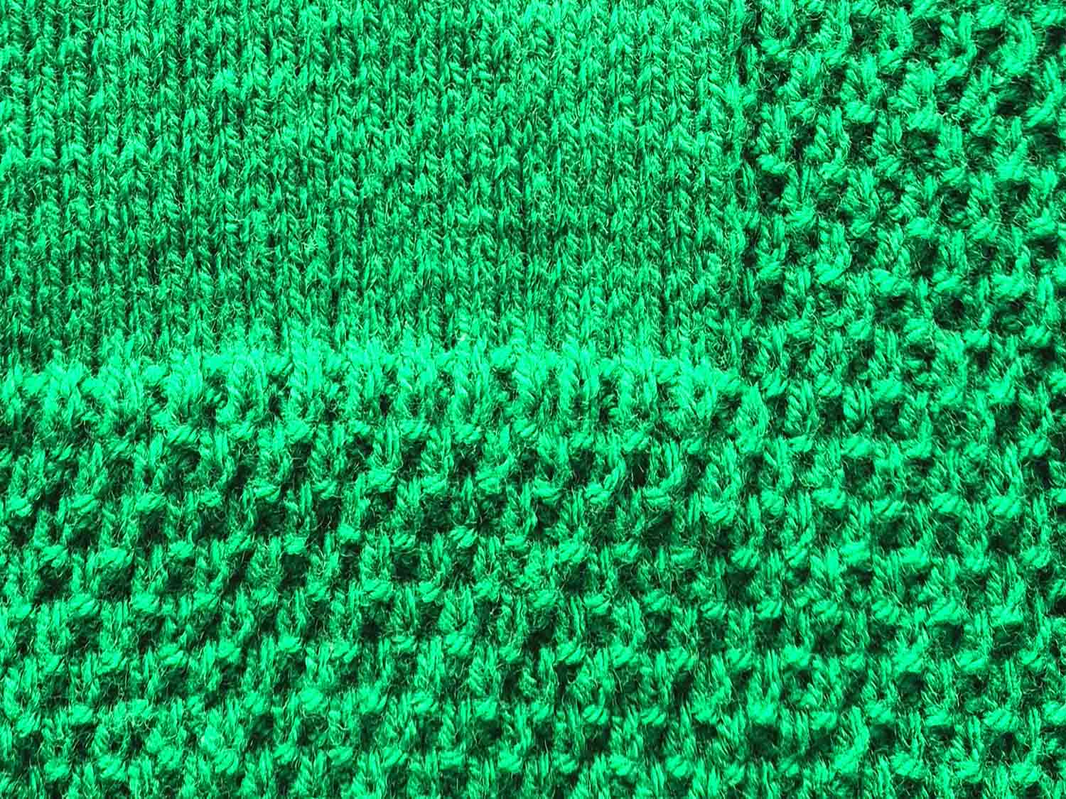 Ein aus grüner Wolle gestrickter Deckenrand im großen Perlmuster ist zu sehen.