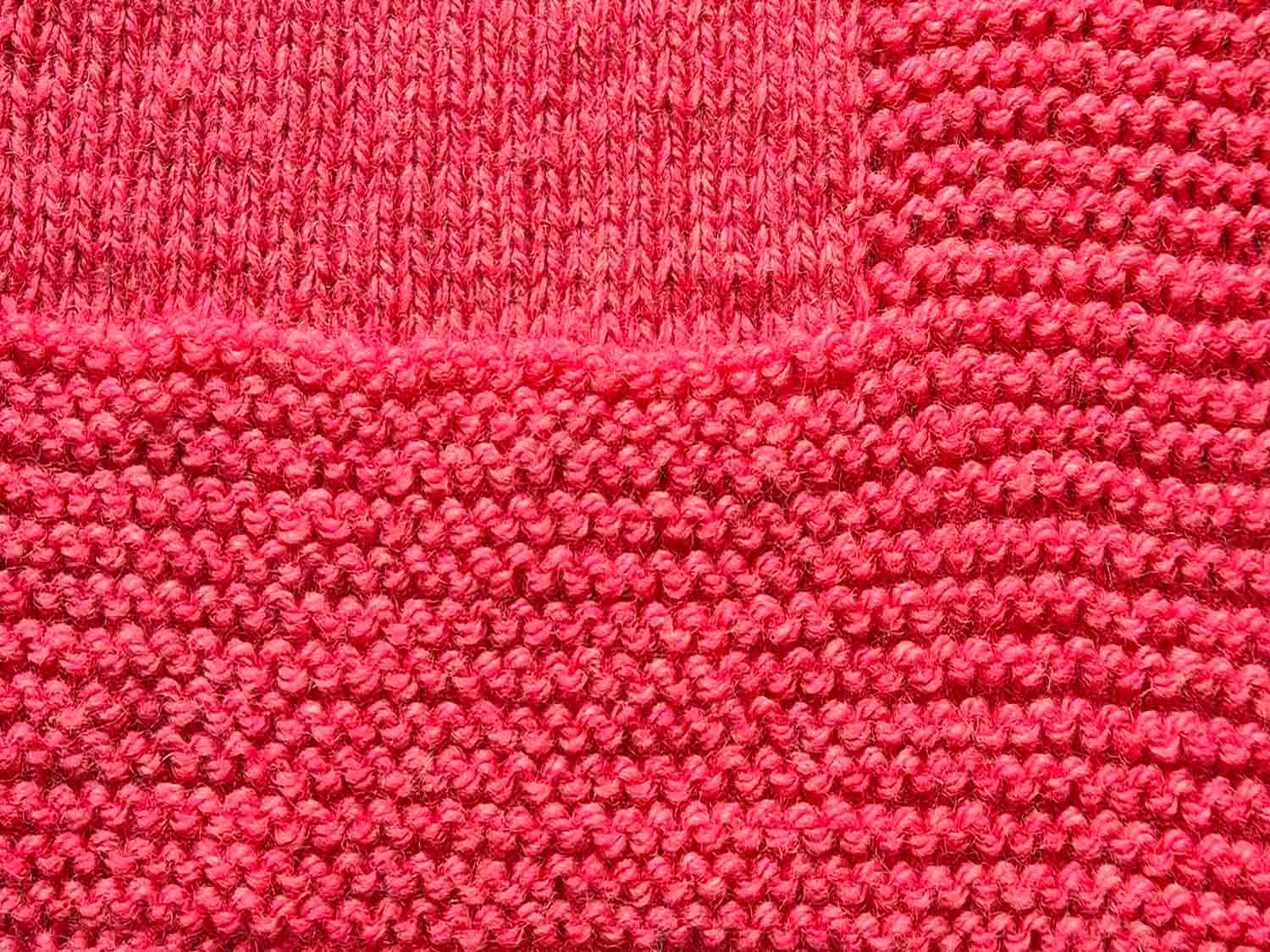 Ein gestrickter Deckenrand aus roter Wolle mit kraus rechts gestricktem Muster ist zu sehen.