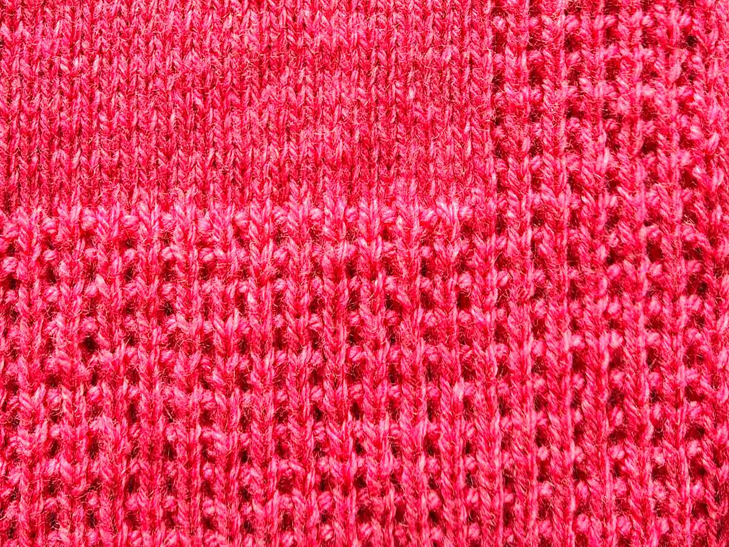 Ein gestrickter Deckenrand mit Längsstreifen und Perlmustersteifen aus roter Wolle ist zu sehen.