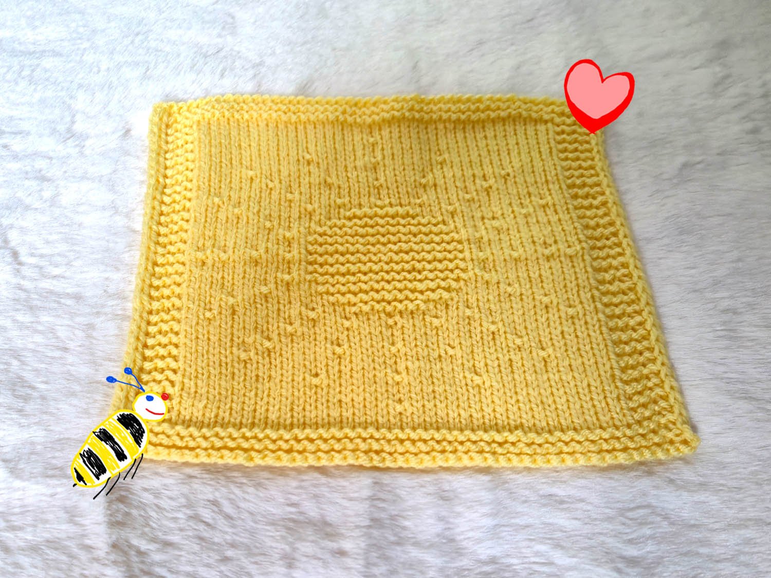 Ein gelbes Tuch mit eingestrickter Sonne liegt auf einem Fell. Ein Herz und eine Biene dekorieren das Sonnentuch.