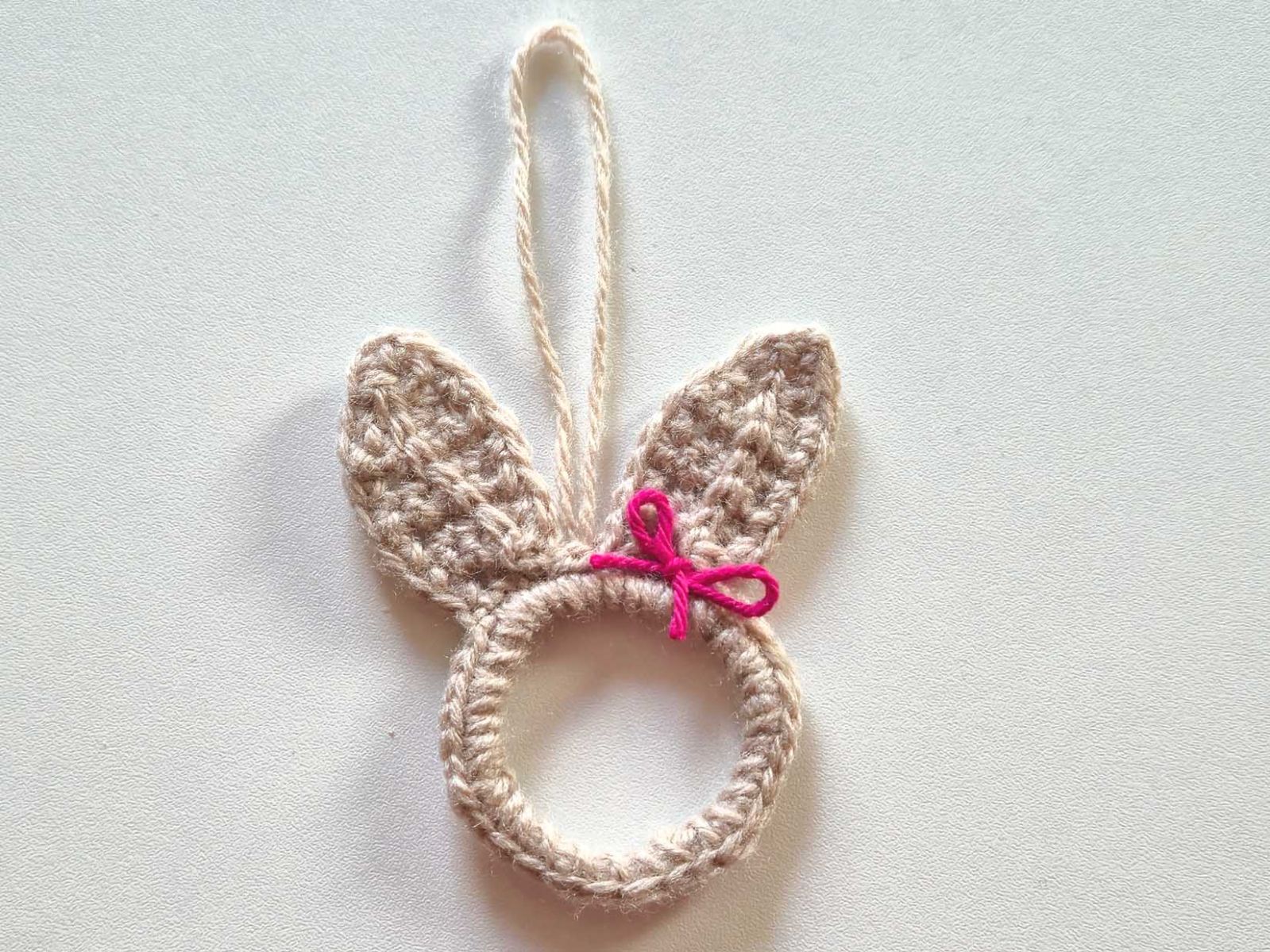 Ein beiger gehäkelter Ring mit Hasenohren als Anhänger dekoriert mit einer pinken Schleife..