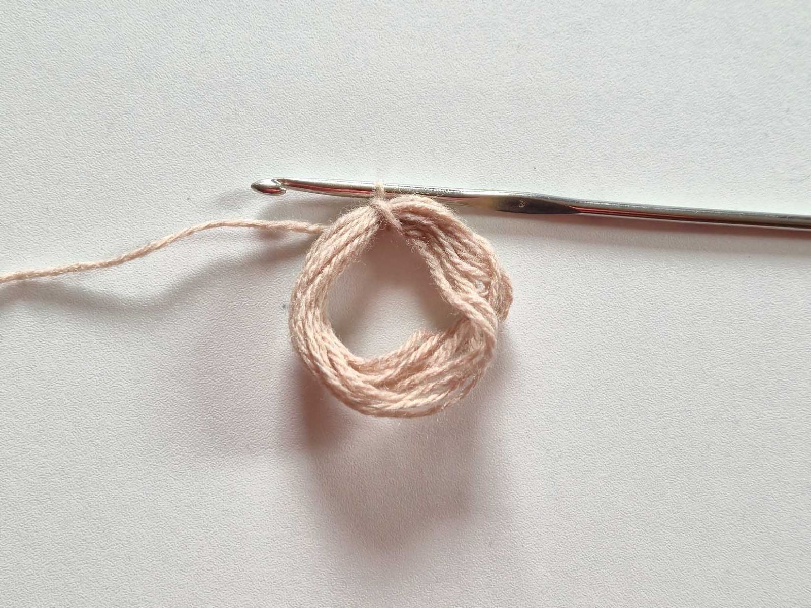 Ein Ring, gewickelt aus mehreren Runden beigem Garn und eine Häkelnadel sind zu sehen.