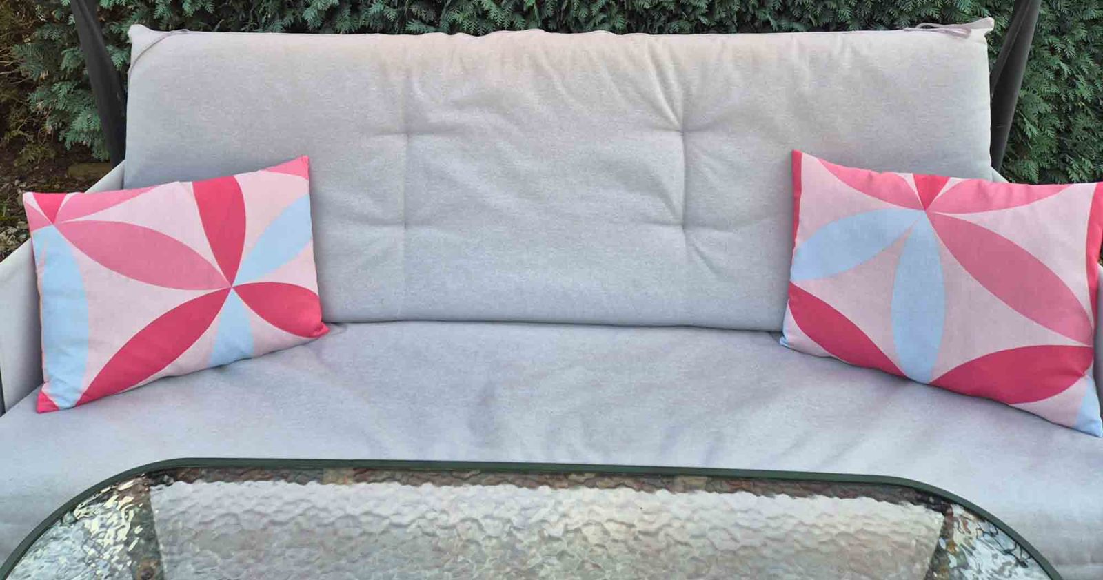 2 dekorative Kissen in verschiedenen Rot- und Rosafarbtönen liegen auf der Sitzfläche einer Hollywoodschaukel.