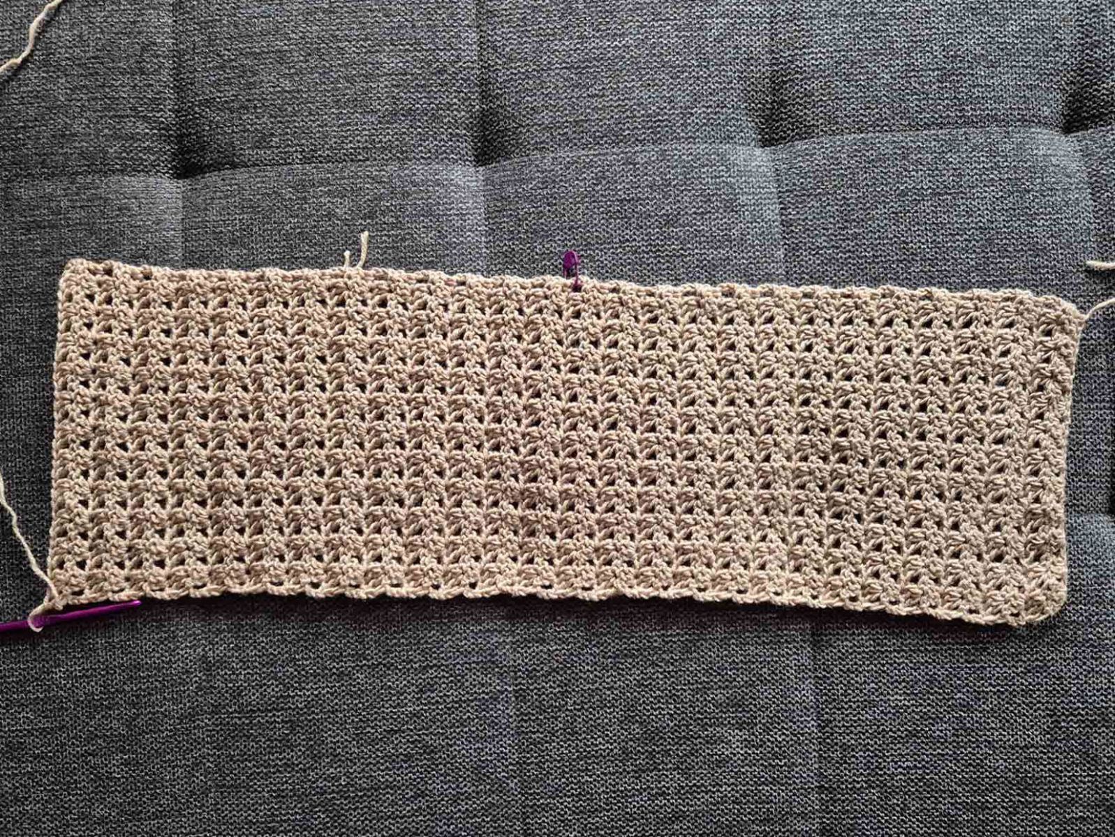 Der als Rechteck gehäkelte beige Schal liegt auf einem dunkelgrauen Untergrund.