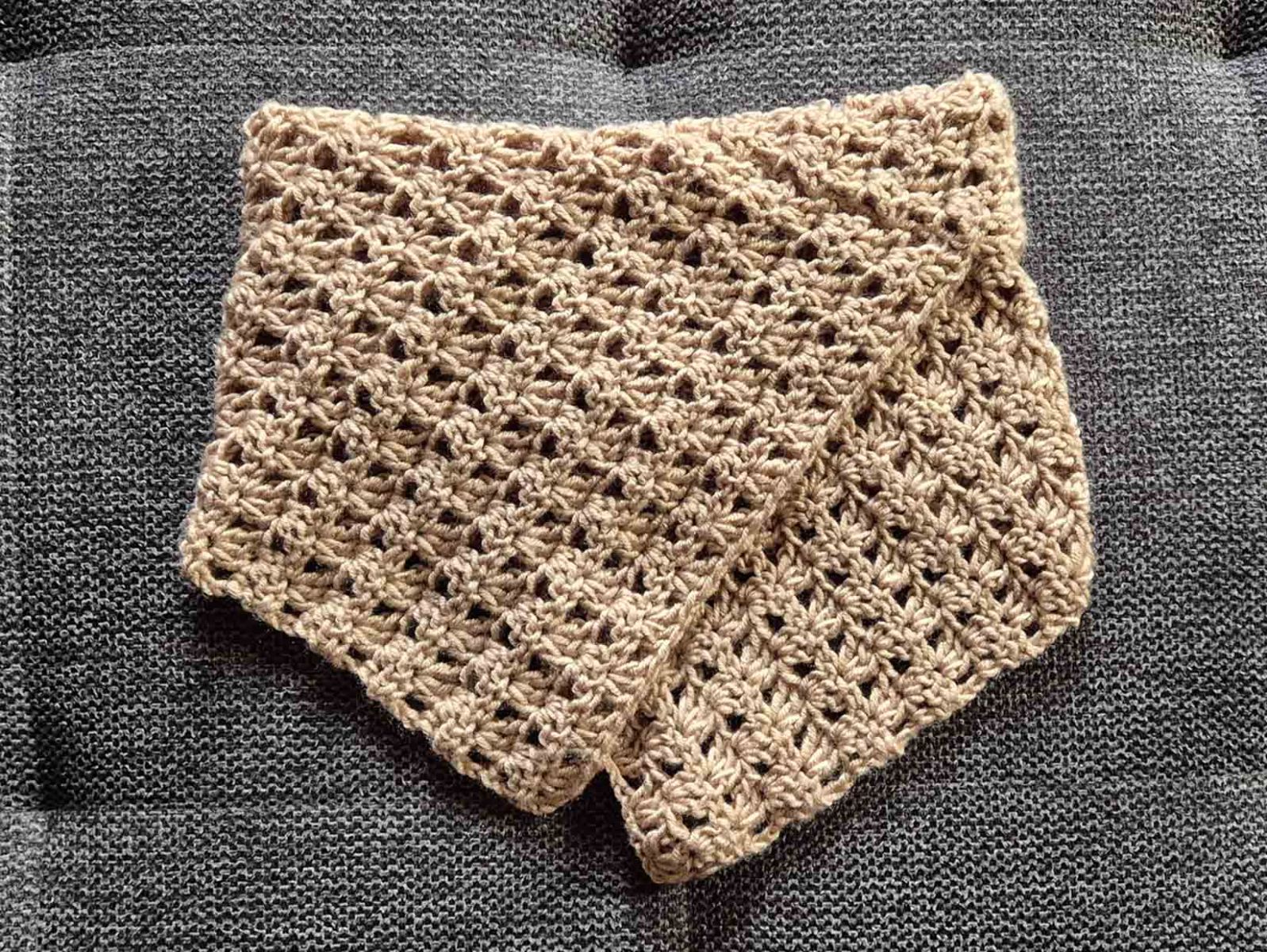 ein gehäkelter Klappschal in beige liegt auf einem grauen Untergrund