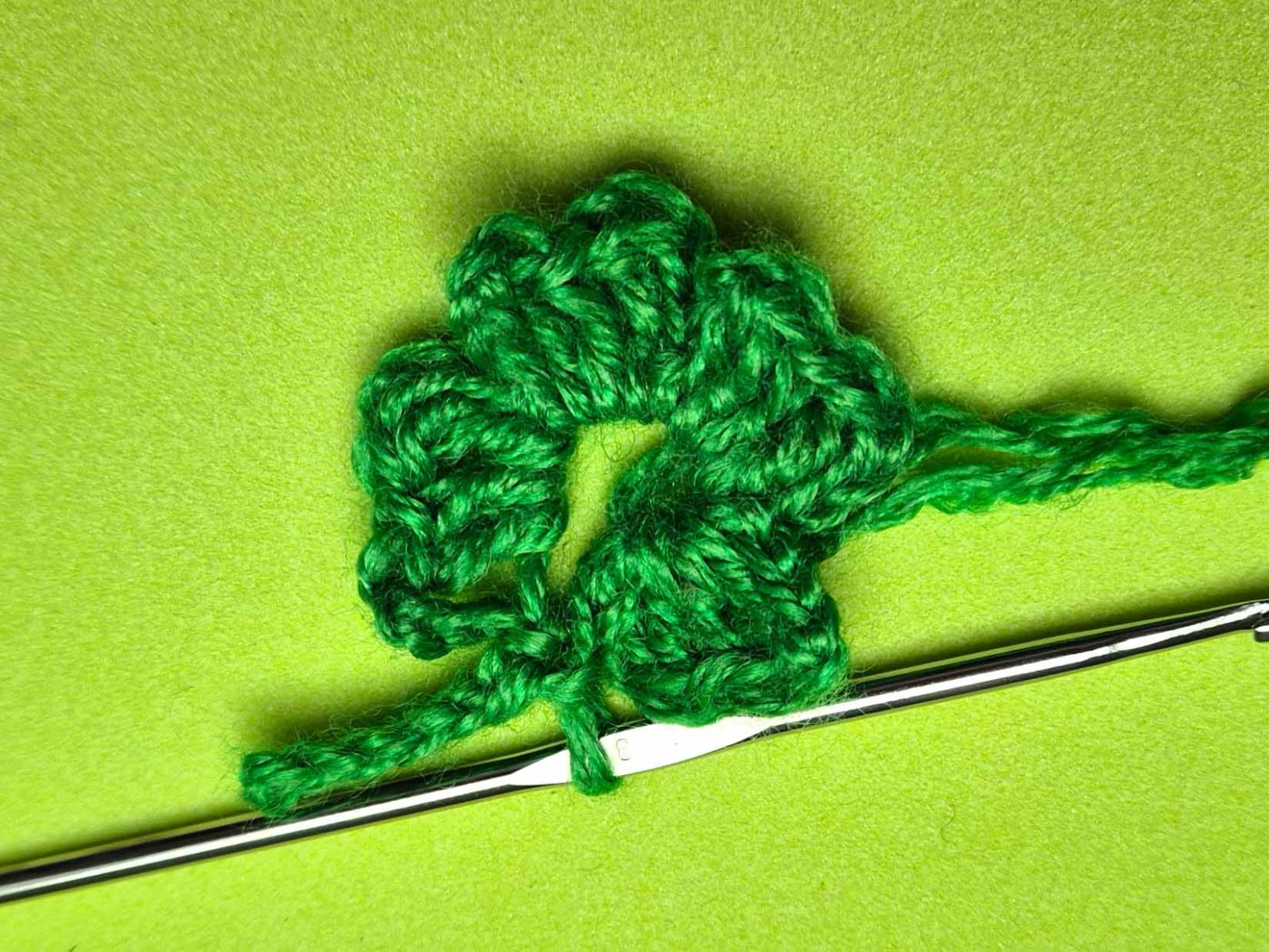 Das gehäkelte Kleeblatt mit Stängel und eine Häkelnadel liegen auf einem grünen Untergrund.