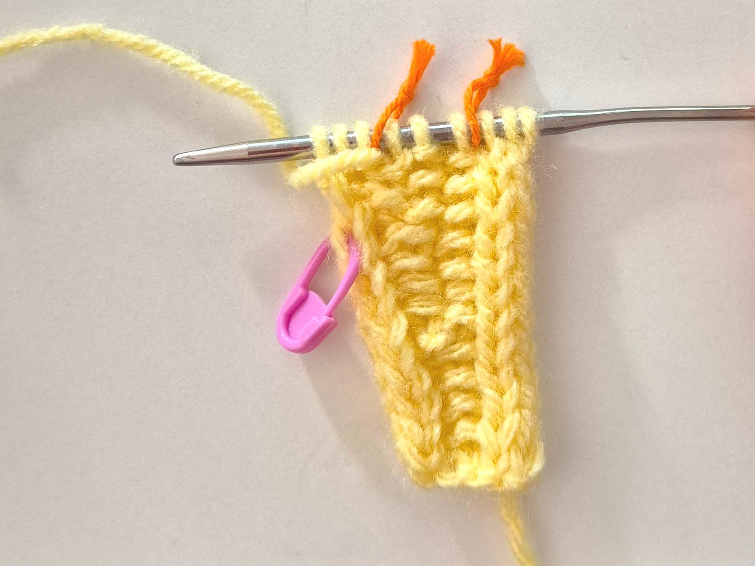 Das gelbe Strickstück wird mit einem Maschenmarkierer links zur Kennzeichnung der Seite für die Zunahmen und mit 2 Maschenmarkierern zur Abtrennung von Rand zu Perlmustermitte gezeicht.