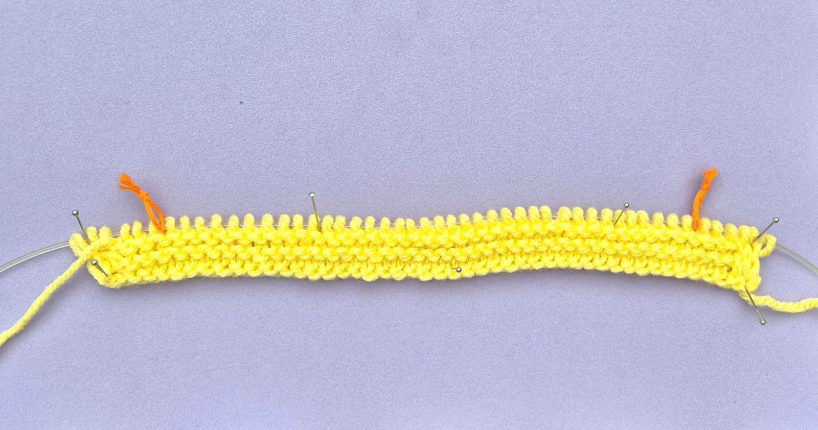 Auf einem fliederfarbigen Untergrund liegt der Anfang des gestrickten Tuchs. 2 orange Maschenmarkierer trennen die Ränder vom Motivteil.