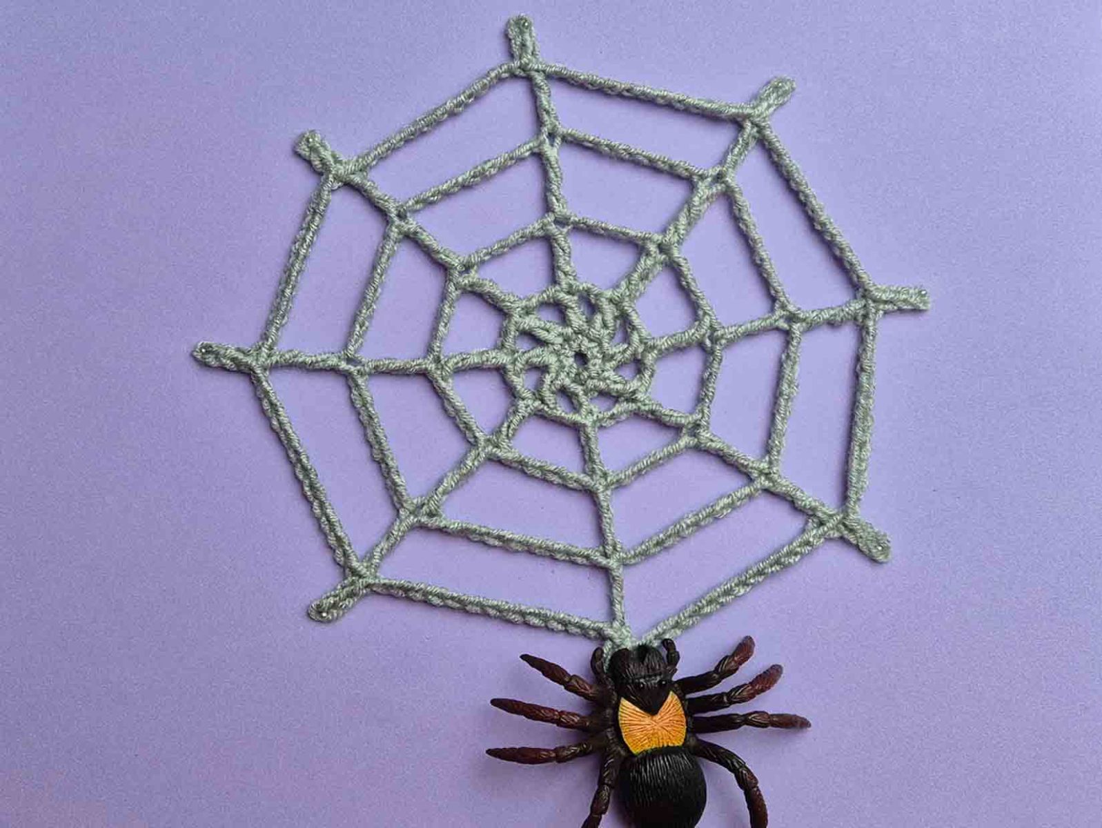 ein graues gehäkeltes Spinnenetz mit einer Spinne liegt auf einem lila Untergrund