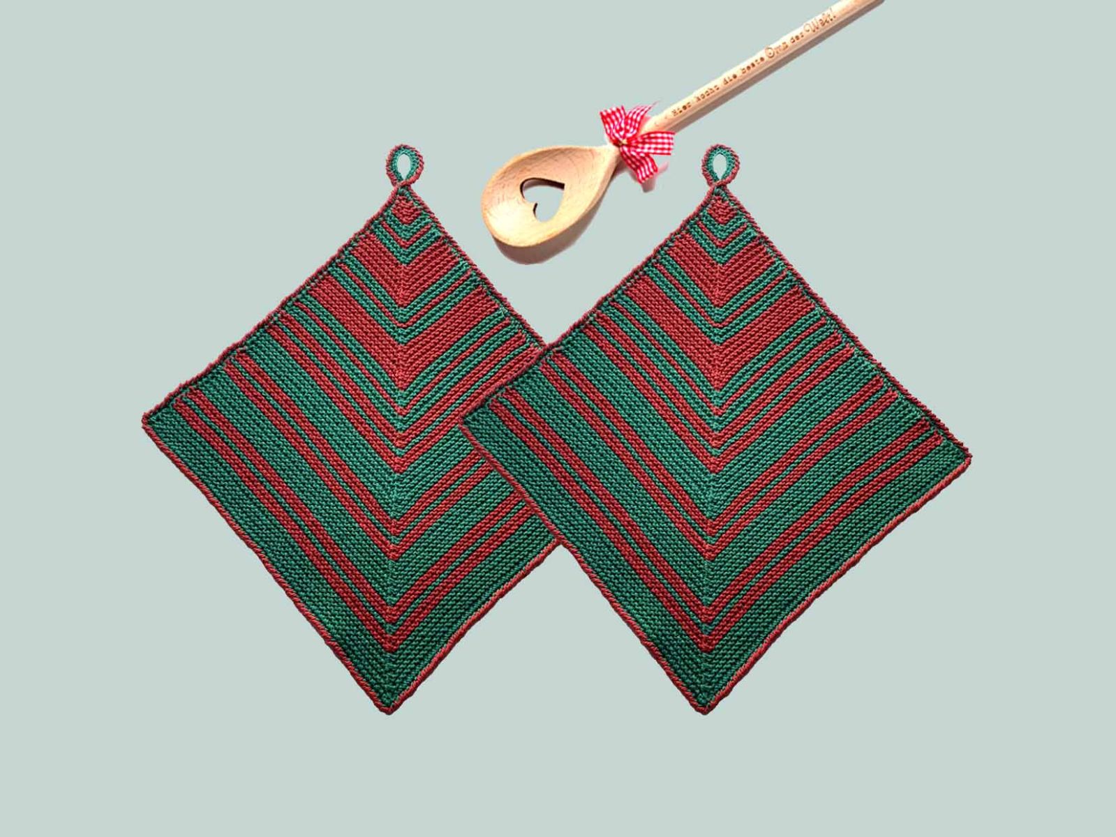Zwei gestrickte Topflappen in grün und weinrot sowie ein Kochlöffel mit rosa Schleife liegen auf einem graugrünen Untergrund.