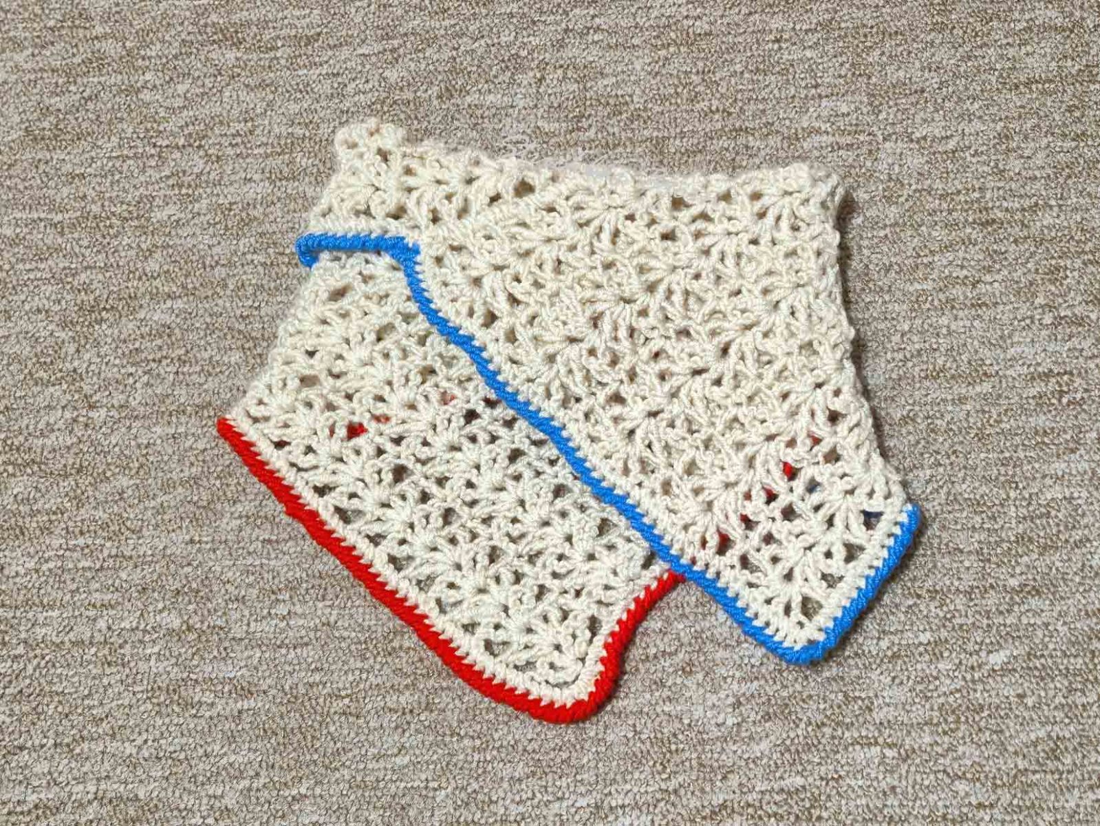 Ein Klappschal in cremeweiß mit rotem und blauem Rand liegt auf einem beigen Untergrund.