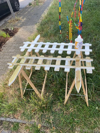 Hier sieht man im Hintergrund die Wind-Spiralen.
Die Zaunelemente werden zunächst weiß grundiert und dann in den Regenbogenfarben lackiert. Und dann kommen sie rings um den Vorgarten.
Das nächste ist eine ebenfalls mit Holz in Regenbogenfarben eingefasste Kräuterspirale, die auch in den Vorgarten gebaut wird.
