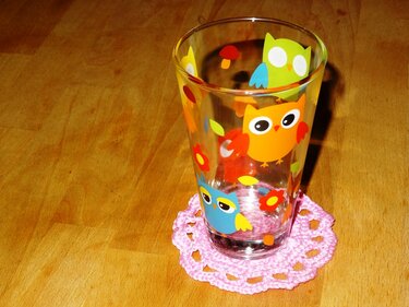 Runder rosa gehäkelter Untersetzer unter einem Glas mit Eulenmotiv auf einem Holztisch.