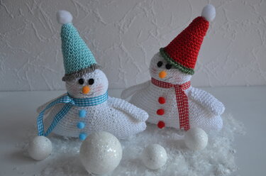 Zwei handgehäkelte Schneemänner mit blauer und roter Mütze sowie karierten Schals auf Dekoschnee neben glitzernden Kugeln.