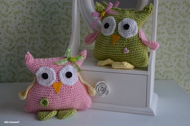 Zwei gehäkelte Amigurumi-Eulen (rosa und grün) mit großen Augen, Schleifen und Knopfapplikationen auf weißer Kommode vor gemustertem Hintergrund.