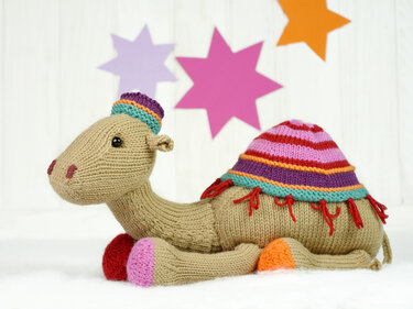 Hand-knitted light brown camel amigurumi with a colorful striped saddle and small hat on a white surface with star-shaped cutouts in the background