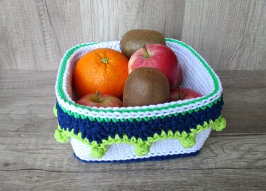 Gehäkelter Korb in Weiß, Dunkelblau und Limettengrün, gefüllt mit Orangen, Äpfeln und Kiwis auf hellem Holztisch.