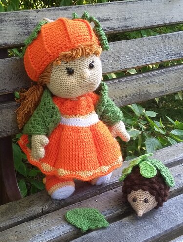 Gehäkelte Amigurumi-Puppe mit orangener Kürbis-Mütze und Kleid, daneben ein kleiner gehäkelter Igel mit grünem Blatt