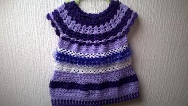 Gehäkeltes Babykleid in Violett, Helllila und Weiß mit Loch- und Streifenmustern, an einem Kleiderbügel hängend