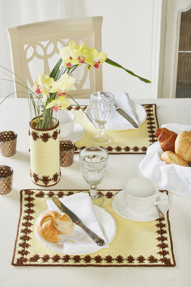 Weißer Esstisch mit zwei gelb-braunen gehäkelten Platzsets, gehäkelter Vasenhülle mit Orchideen, Gläser, Tasse und Brötchen