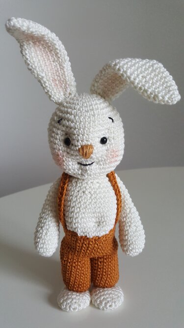 Weißer gehäkelter Hase mit rosa Wangen und brauner Latzhose, stehend