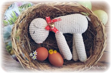 Beiges gehäkeltes Osterlamm mit roter Schleife und Glöckchen in einem Strohkorb neben zwei braunen, mit Blüten verzierten Eiern.