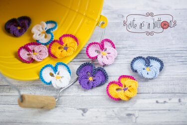 Mehrere kleine gehäkelte Stiefmütterchen-Applikationen in Gelb, Lila, Blau und Rosa auf hellem Holzhintergrund neben einem gelben Eimer