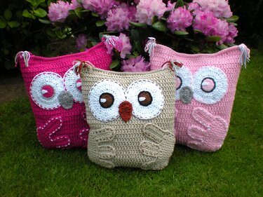 Drei gehäkelte Eulen-Kissenbezüge in Beige, Pink und Rosa auf grünem Rasen vor rosa Rhododendronblüten