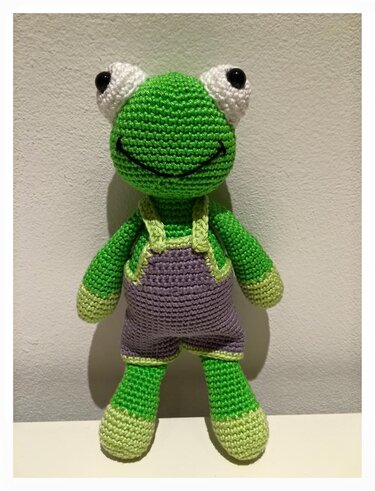 Grüner Amigurumi-Frosch mit lila Latzhose, aufrecht stehend