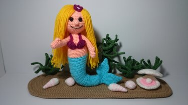 Handgehäkelte Meerjungfrau-Amigurumi mit gelbem Haar, türkisfarbenem Schwanz, Muscheln und grünem Seegras auf einer braunen Häkelunterlage