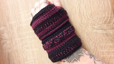 Hand mit gehäkelter Armstulpe in Schwarz und Weinrot auf Holzuntergrund