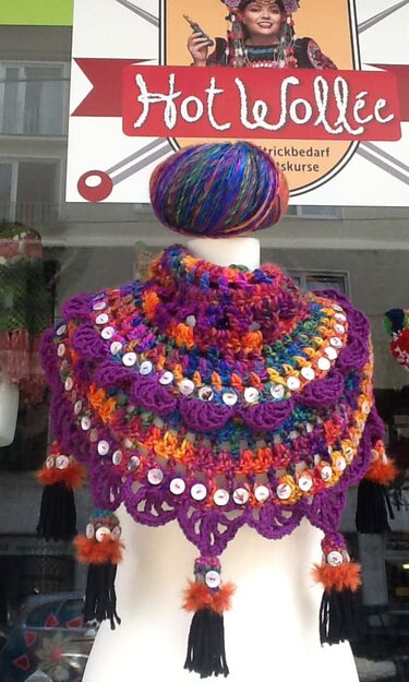 Mannequin trägt einen gehäkelten, mehrfarbigen Kragenschal mit weißen Knöpfen und Quasten