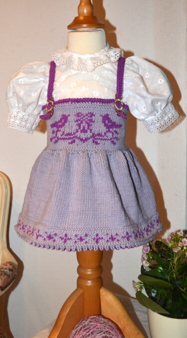Lila gestricktes Baby-Trägerkleid mit Vögelmuster über weißer Spitzenbluse auf Holzpuppe