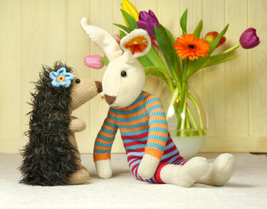 Knitted hedgehog with blue flower facing striped amigurumi rabbit, tulips in vase behind