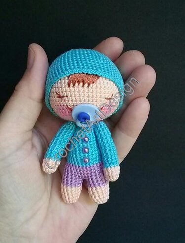 In einer Hand gehaltenes, gehäkeltes Amigurumi-Baby mit blauer Mütze und Schnuller.