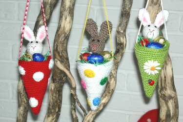 Drei gehäkelte konische Hängekörbchen in Rot, Weiß und Grün mit kleinen Häkelhasen und bunten Eiern, an naturbelassenen Ästen aufgehängt.