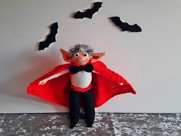 Gehäkelte Vampirpuppe mit rotem Umhang und schwarzer Fliege vor weißer Wand mit Fledermaus-Dekor
