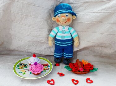 Gehäkelte Jungenpuppe in blau-weißem Outfit, umgeben von Cupcakes, Rosen, herzförmigen Dekorationen und einem Marienkäfer