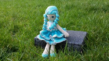 Gehäkelte Amigurumi-Puppe in türkis-weißem Rüschenkleid mit gedrehten Zöpfen, sitzend auf Stein im Gras