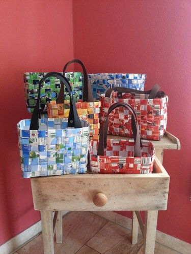 Handgeflochtene Taschen aus recycelten Getränkekartons in verschiedenen Farben auf einem Holztisch vor roter Wand