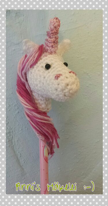 Gehäkelter weißer Einhornkopf mit rosa Mähne und schimmerndem Horn als Stiftaufsatz auf rosa Stift.