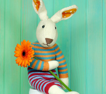 Knitted Easter bunny amigurumi in bright striped outfit holding an orange flower against a teal wooden backdrop
