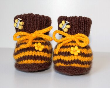 Zwei handgestrickte Babyschuhe in Braun und Gelb mit Streifen, gelben Schleifen und Blüten- sowie Bienenknöpfen.