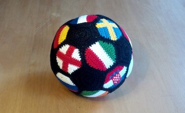 Handgehäkelter Ball mit bunten Länderflaggen auf dunklem Grund, auf Holzuntergrund liegend.
