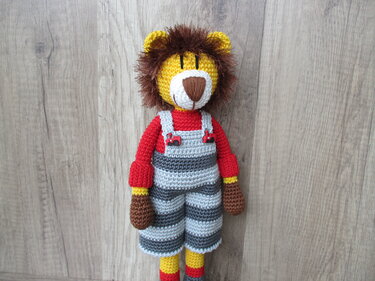 Gehäkelte Löwenpuppe mit brauner Mähne, rotem Shirt und grau-dunkelgrau gestreifter Latzhose vor Holzuntergrund