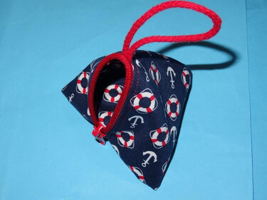 Dreieckige dunkelblaue Stofftasche mit Anker- und Rettungsring-Motiven, rotem Reißverschluss und roter Trageschlaufe auf hellblauem Hintergrund.