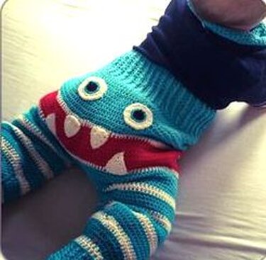 Nahaufnahme einer gehäkelten türkis-weiß gestreiften Babyhose mit rotem Monstermund und aufgenähten Augen.