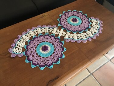 Gehäkelter Tischläufer aus zwei runden Blumenmotiven in Lila, Türkis und Creme auf Holztisch